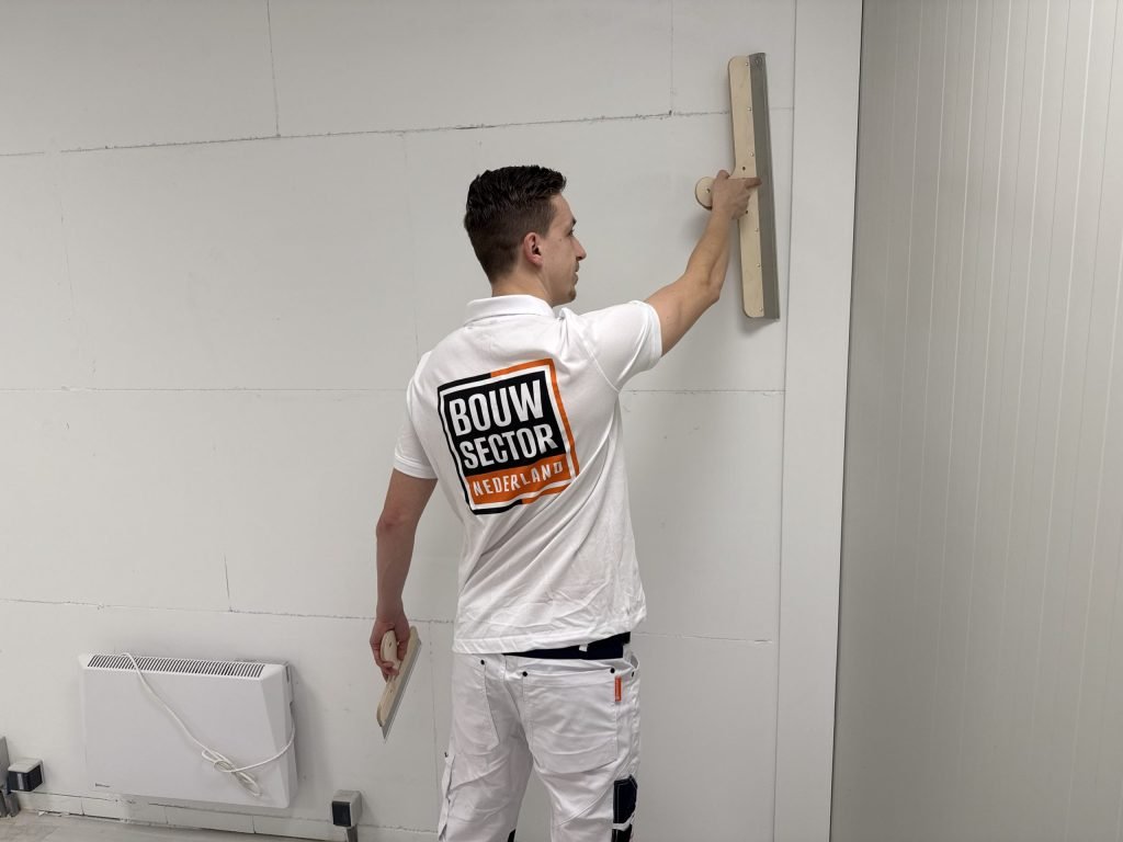 Stukadoor van Bouwsector Nederland trekt met een lange spaan de bovenkant van een wand vlak, naast een kachel