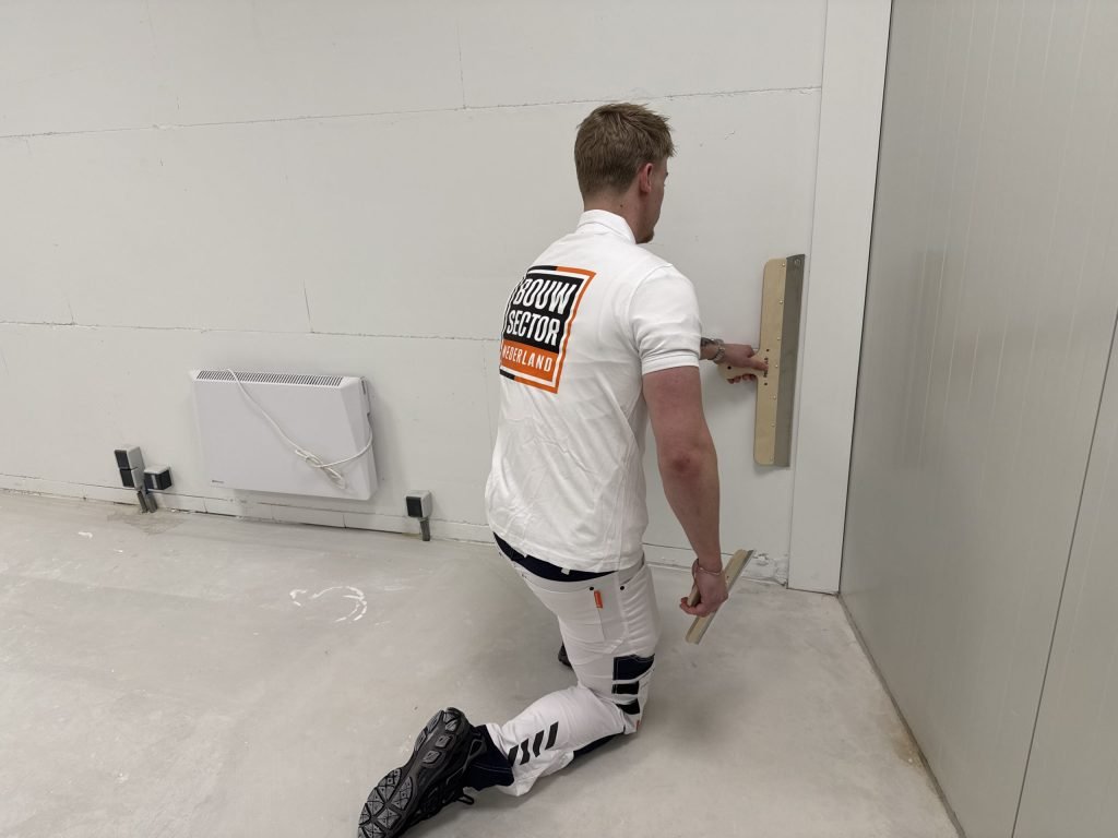 Stukadoor op een knie zet de onderkant van een wand vlak met een spaan, vlak boven de vloer