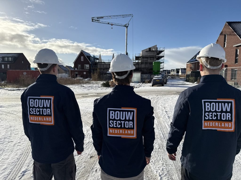Drie vakmensen van Bouwsector Nederland met witte helm voor een besneeuwde nieuwbouwstraat met een bouwkraan